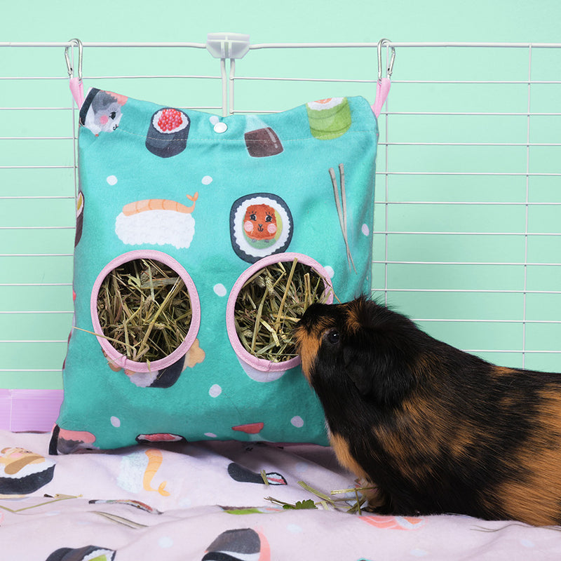 Black and brown guinea pig eating hay from Kavee limited edition sushi design haybag inside white cage on sushi design fleece liner