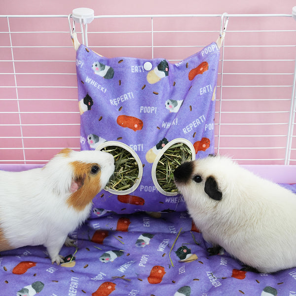 Two guinea pigs eating hay from from purple poop design haybag by brand Kavee in white cage on purple poop design fleece liner