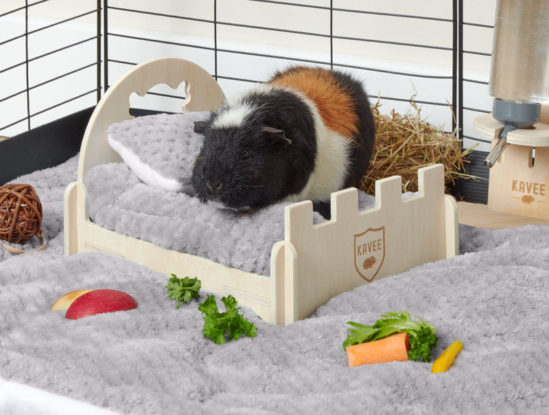 tri coloured guinea pig snuggling next to gray fleece pillow on miniature wooden castle bed with gray pee pad